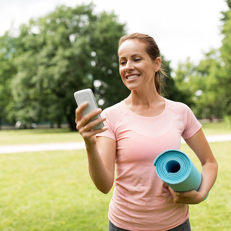 fitness, sport and healthy lifestyle concept - happy smiling woman with exercise mat and smartphone at park