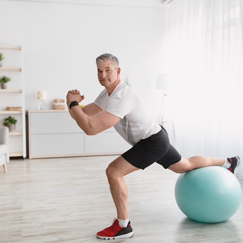 Home workout. Senior man doing lunges with fitball, exercising at home and smiling at camera, copy space. Mature man making domestic training during coronavirus isolation