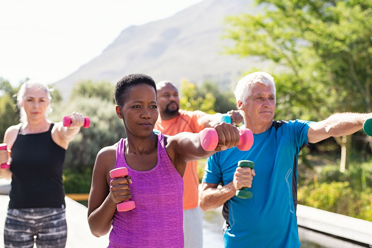 Happy seniors and mature couples exercising with dumbbells. Healthy multiethnic people exercising using dumbbells outdoor. African couple and senior friends in sportswear stretching arms at park.